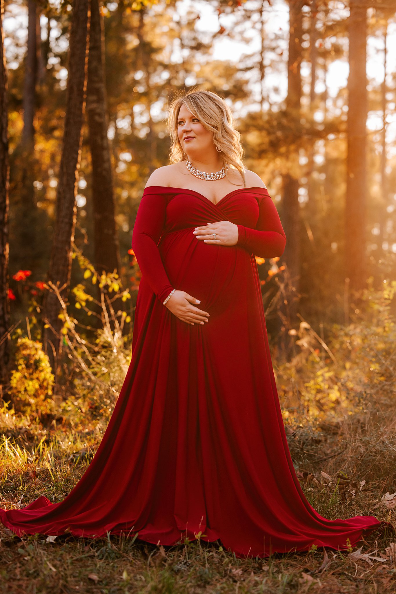 Mother to be holds her bump in a large necklace and red maternity gown Houston Baby Boutique
