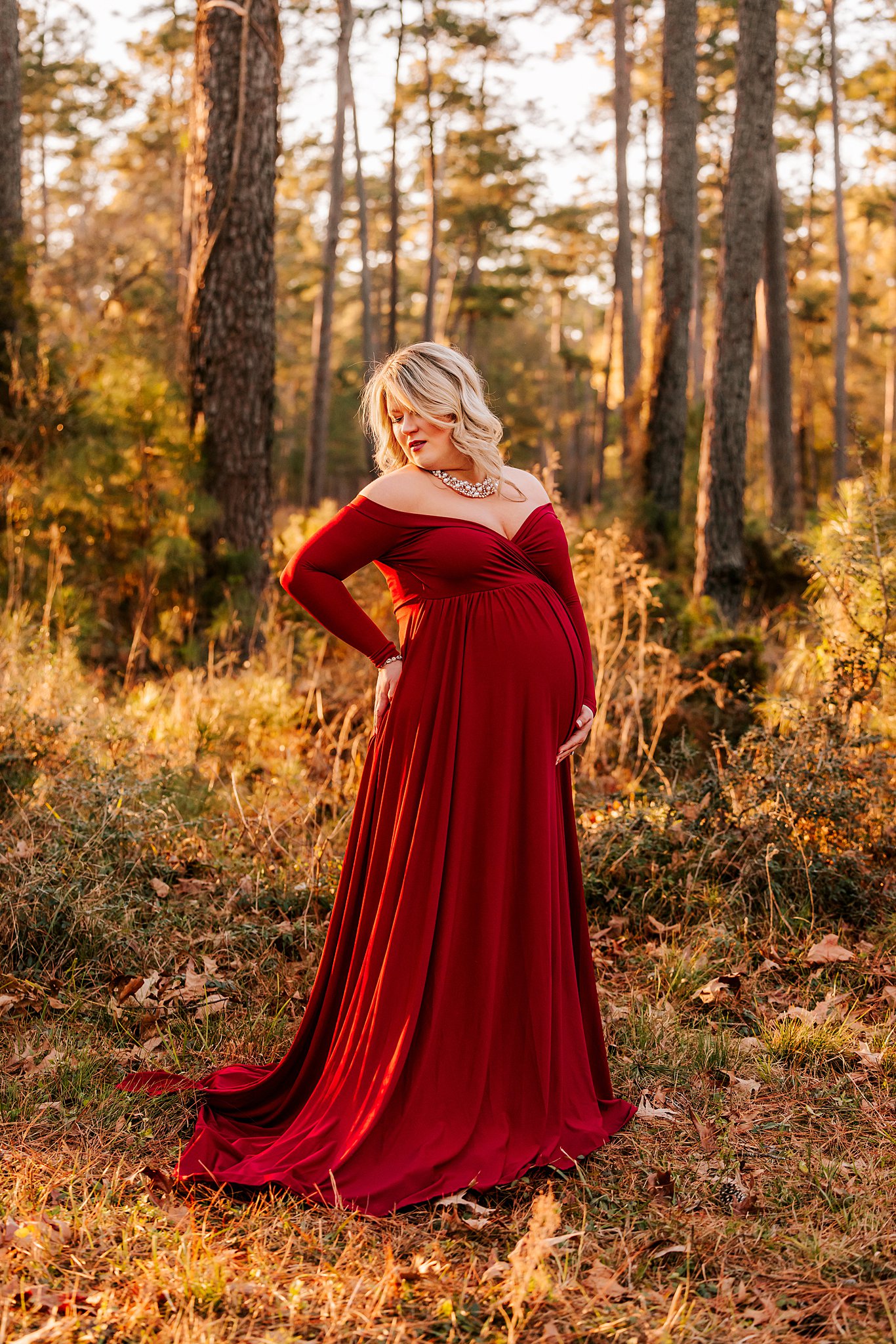 Mother to be stands in the forest holding her bump in a red maternity gown