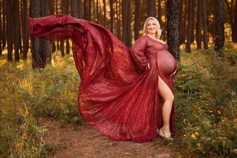 Mom to be stands in the forest with wildflowers and a flowing red maternity gown