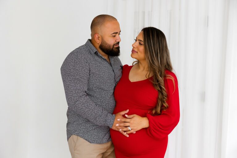 dad and mom to be look at each other in a studio with hands on her bump in a red maternity gown