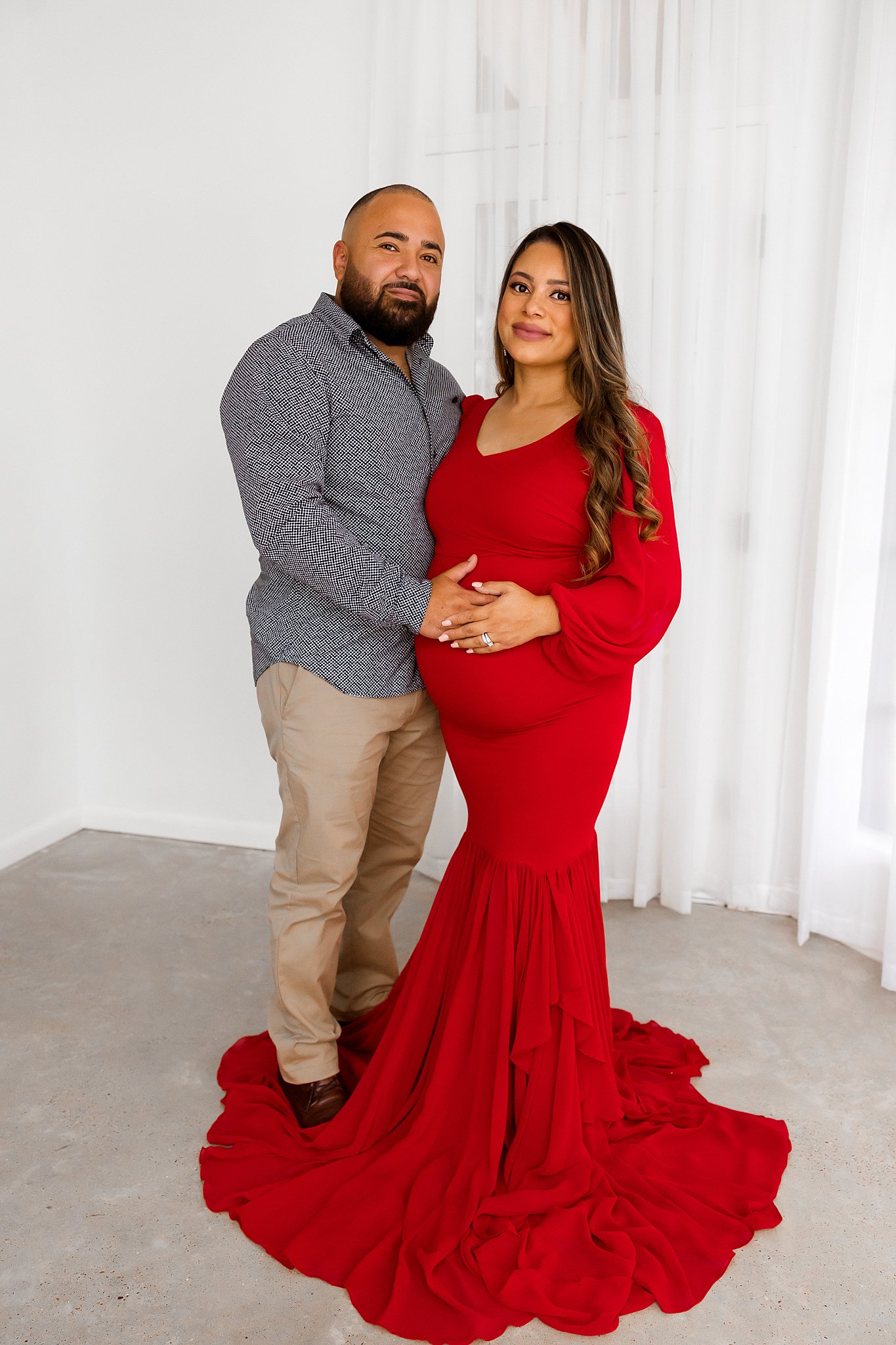 Husband and Mom to be stand in a studio while wearing a read maternity gown houston birth center